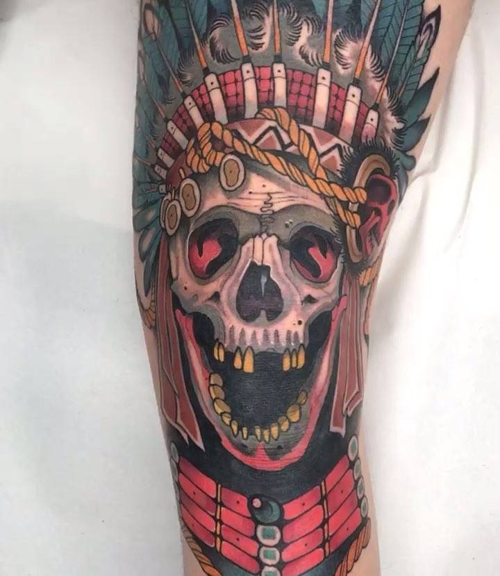 Neotraditional skull and war bonnet tattoo on the knee.