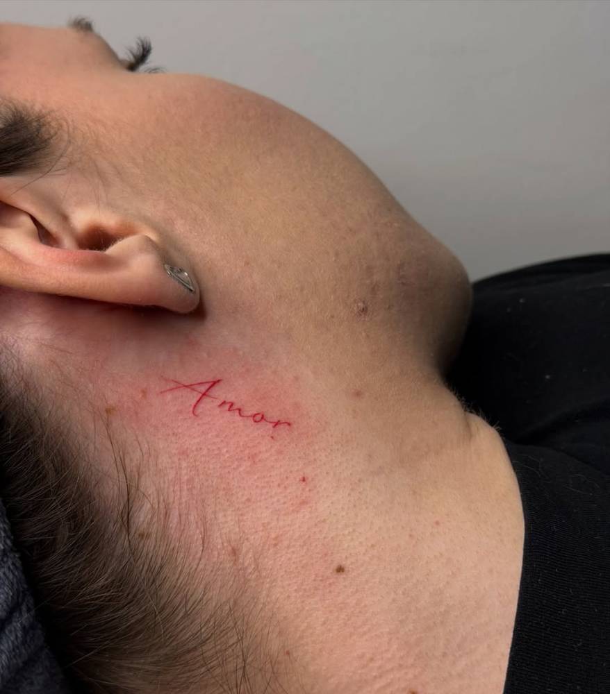 Lettering tattoo of the word "amor" in red ink placed behind the ear.