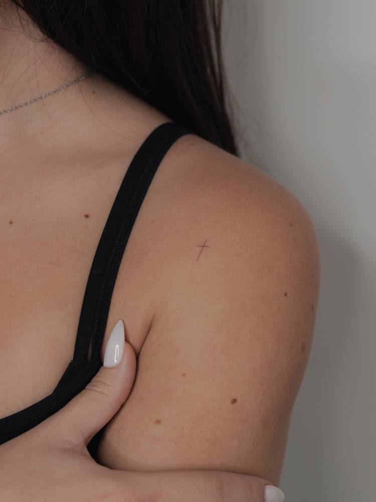 Tiny minimalistic cross tattoo located on the shoulder.
