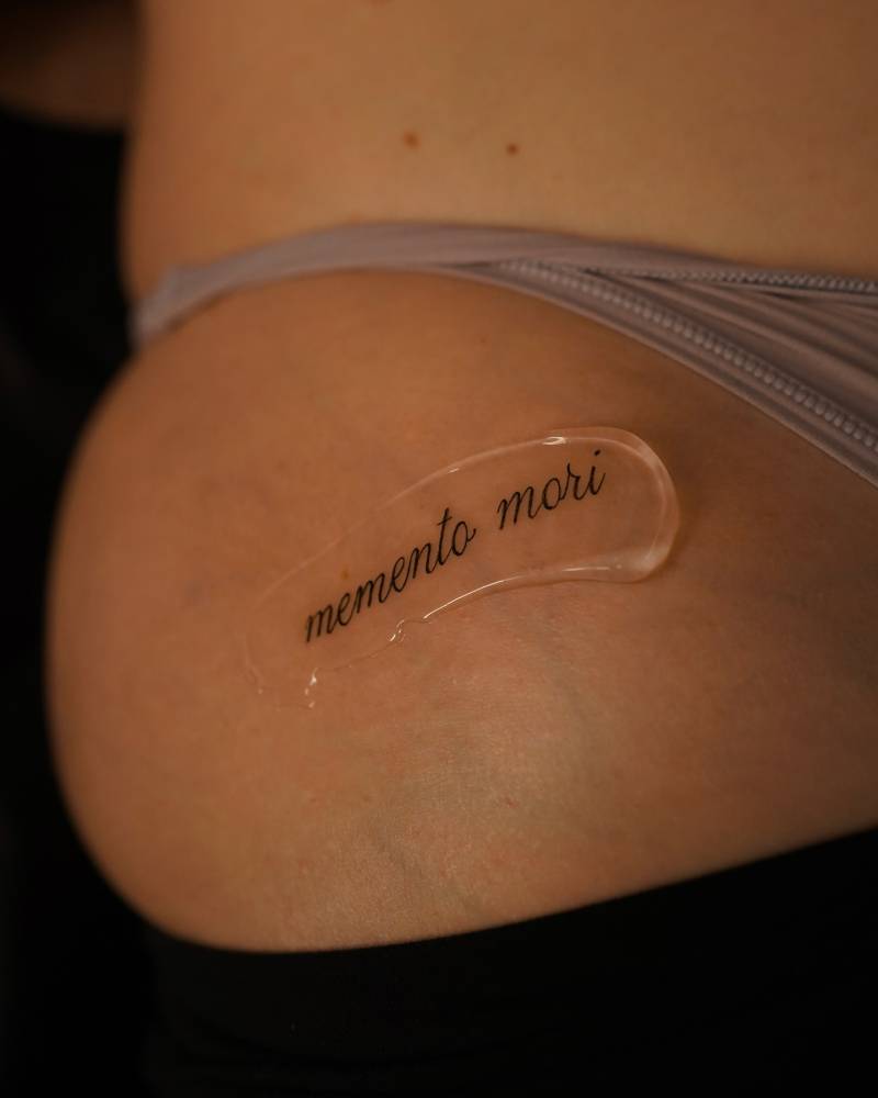Script font "memento mori" lettering tattoo located on the hip.