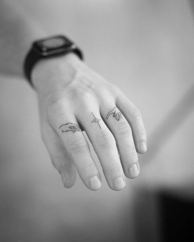 Minimalistic style 'Creation of Adam' hands and star tattoo located on the fingers.