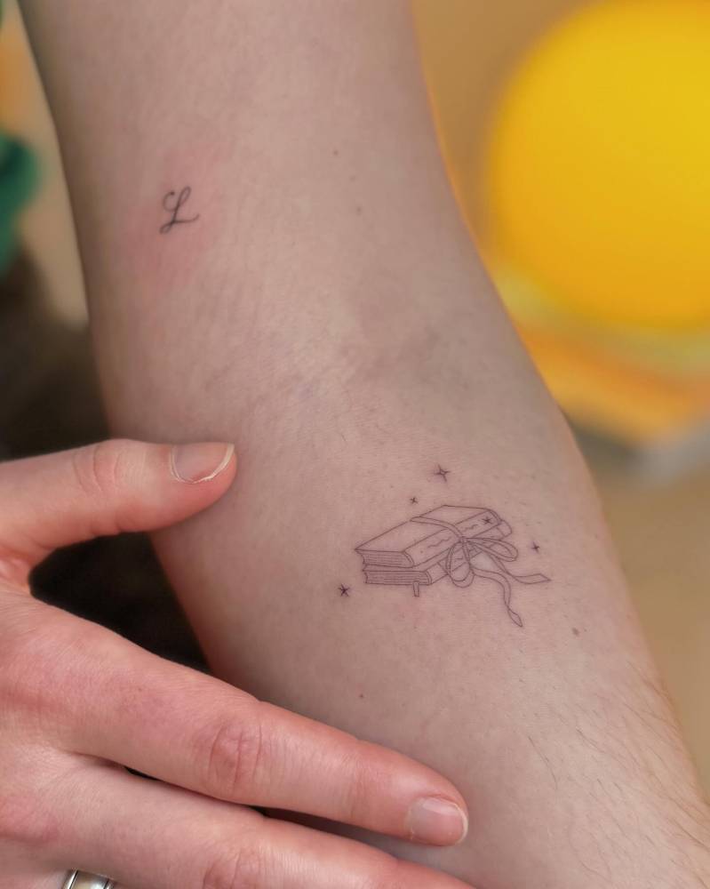 Fine line book and tiny letter "L" tattoos.