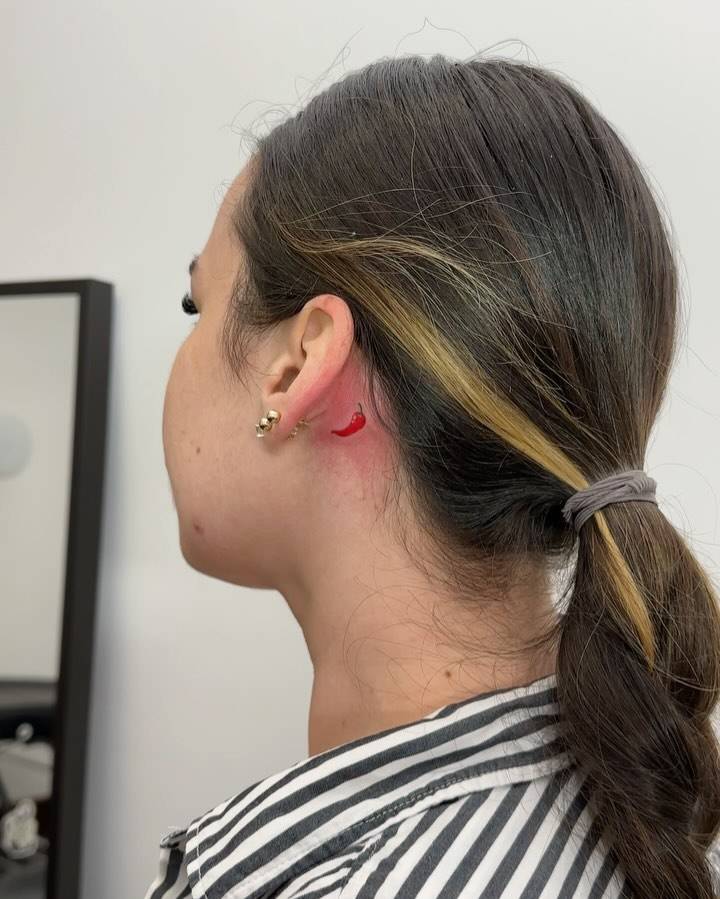 Tiny red chili tattoo placed behind the ear, illustrative style.
