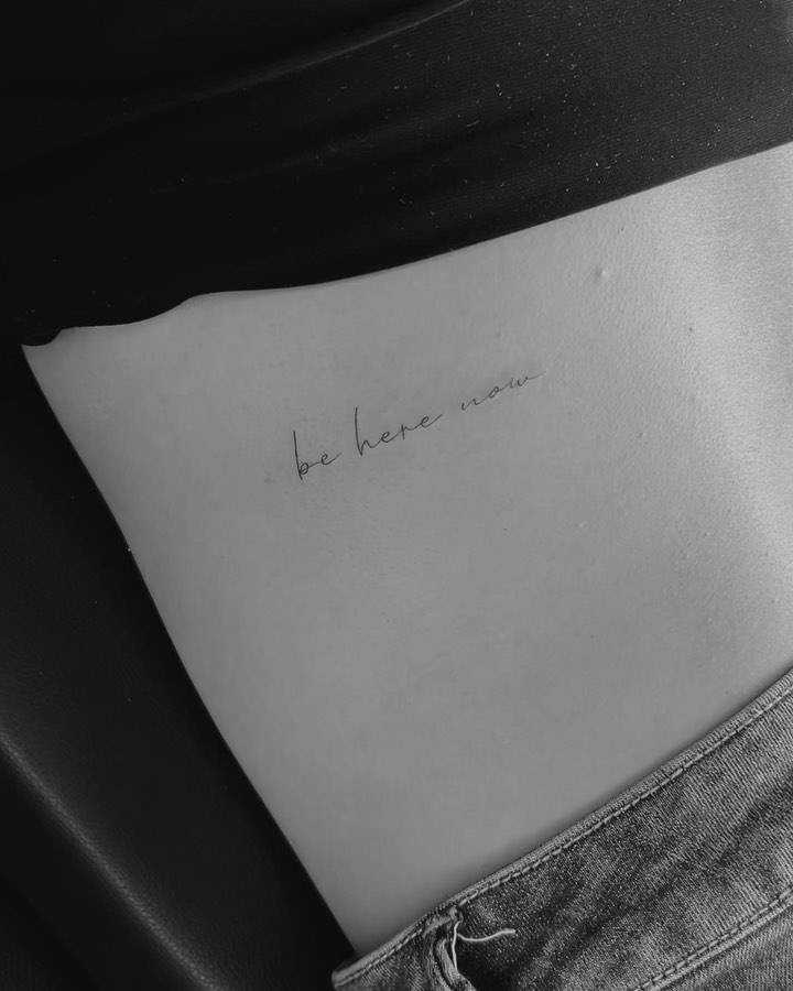 "Be here now" lettering tattoo handwritten on the lower back.