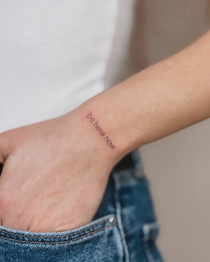 "Be here now" lettering tattoo located on the wrist.