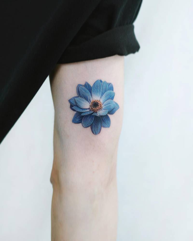 Blue dahlia flower tattooed on the inner arm, micro-realistic style.