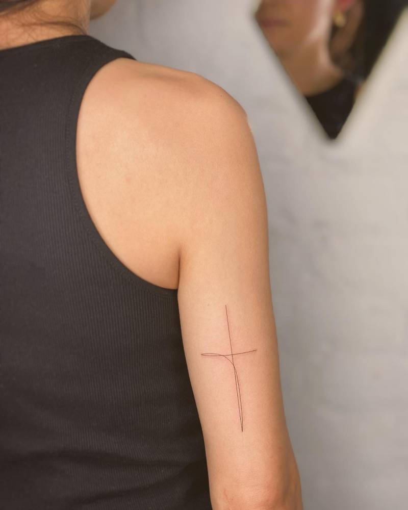 One line cross tattoo in minimalistic style placed on the tricep.