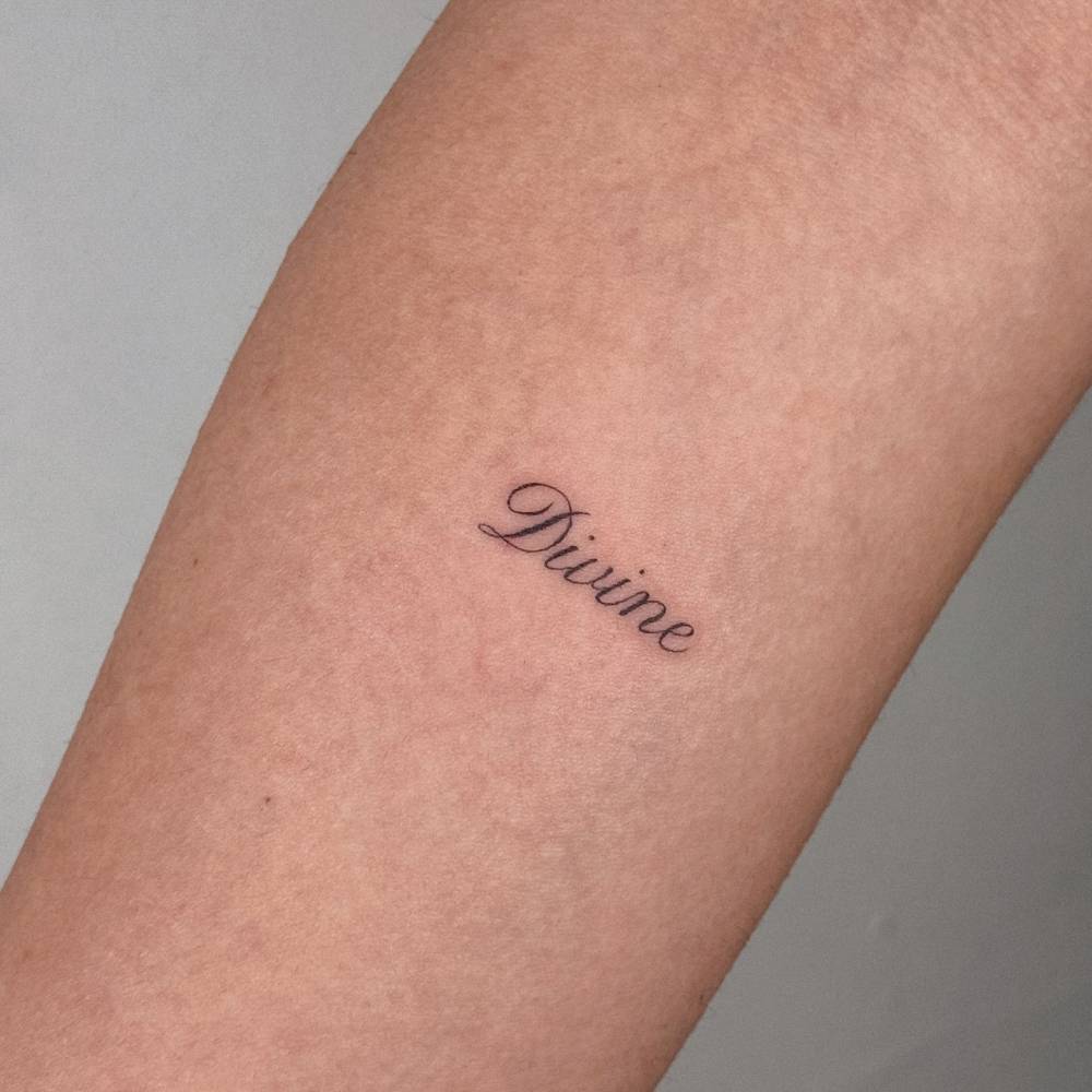 Word "Divine" lettering tattoo located on the inner forearm.