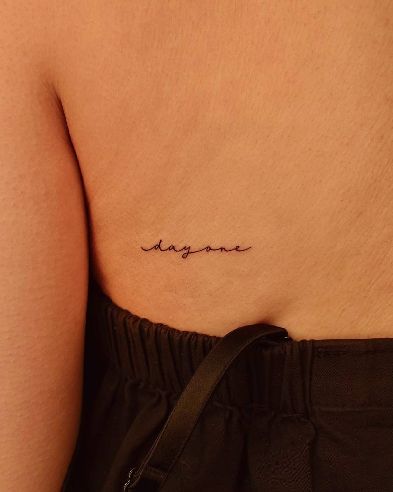 "Day one" handwritten lettering tattoo placed on the back.