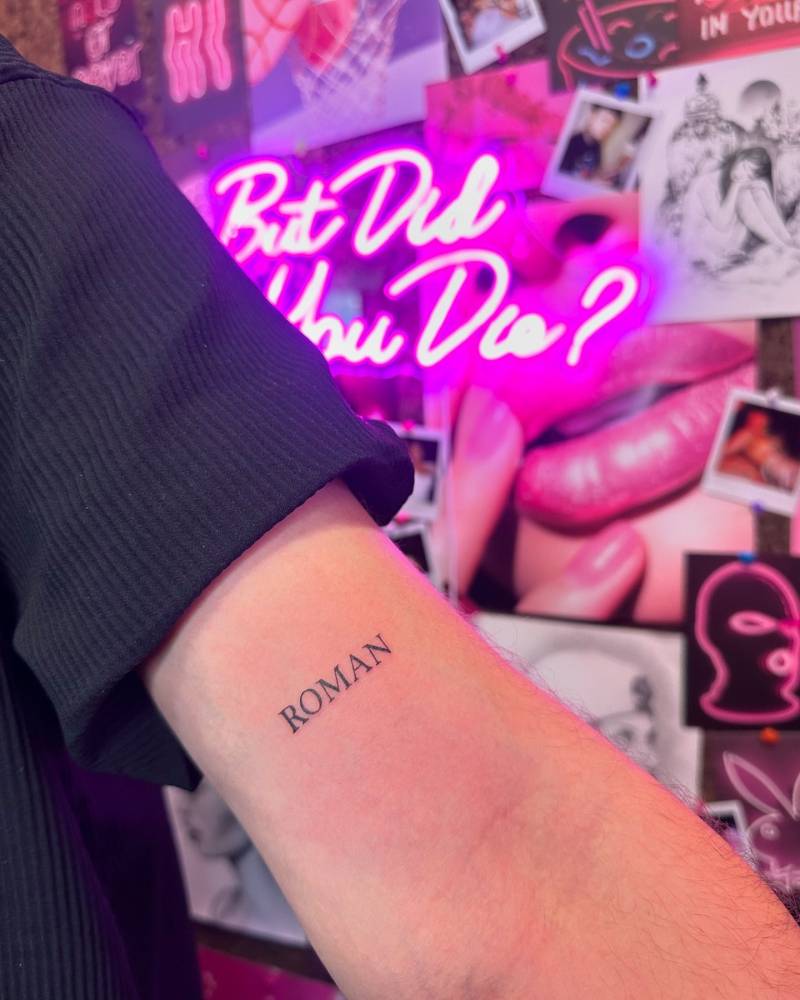 Lettering tattoo of the name "Roman" located on the bicep.