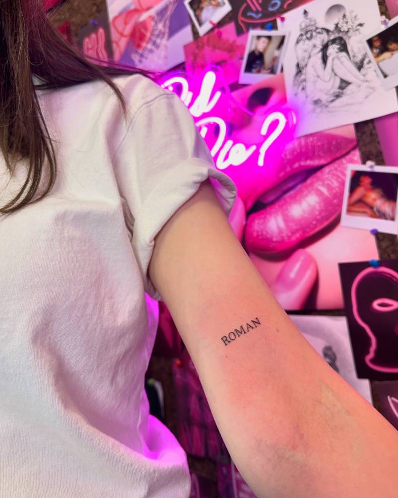 Lettering tattoo of the name "Roman" located on the bicep.
