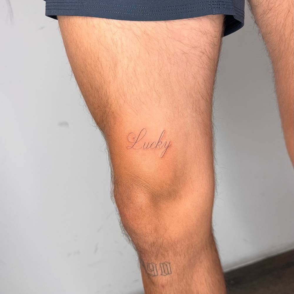 Script font word "lucky" tattooed on the thigh.