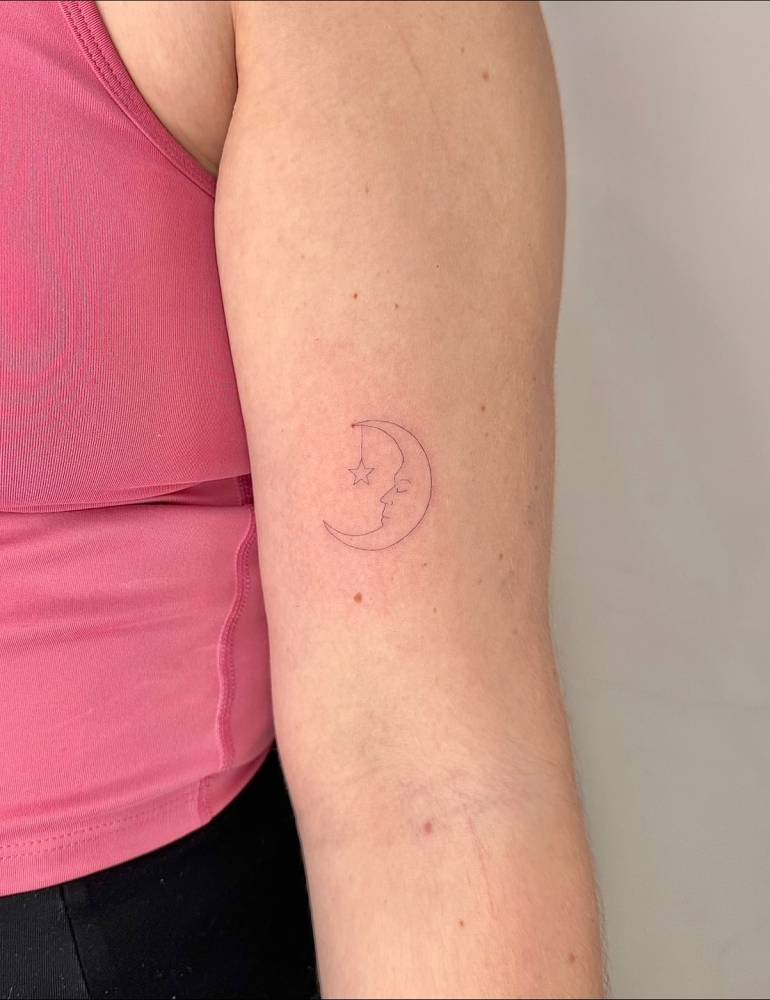 Crescent moon face tattoo located on the bicep, done in fine line style.