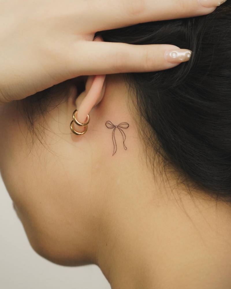 Tiny minimalistic bow tattoo located behind the ear.