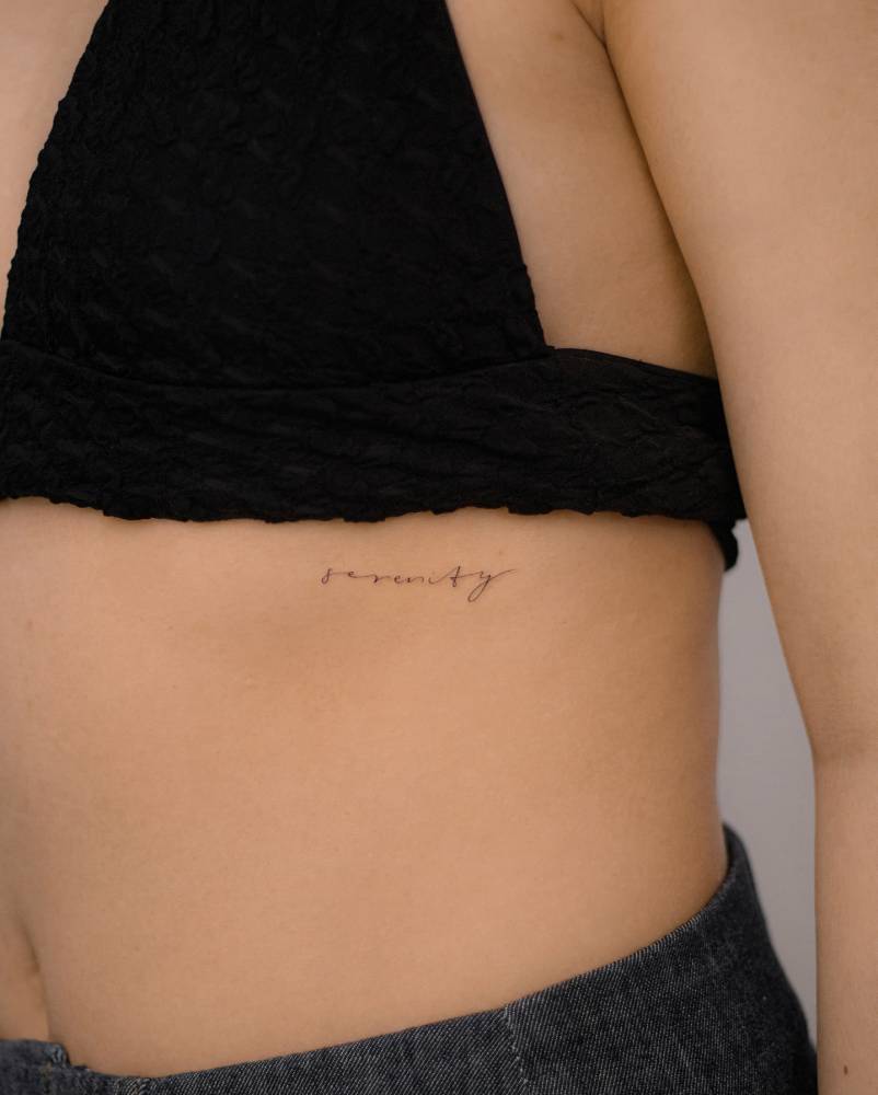 Handwritten font word "serenity" tattoo located on the rib.