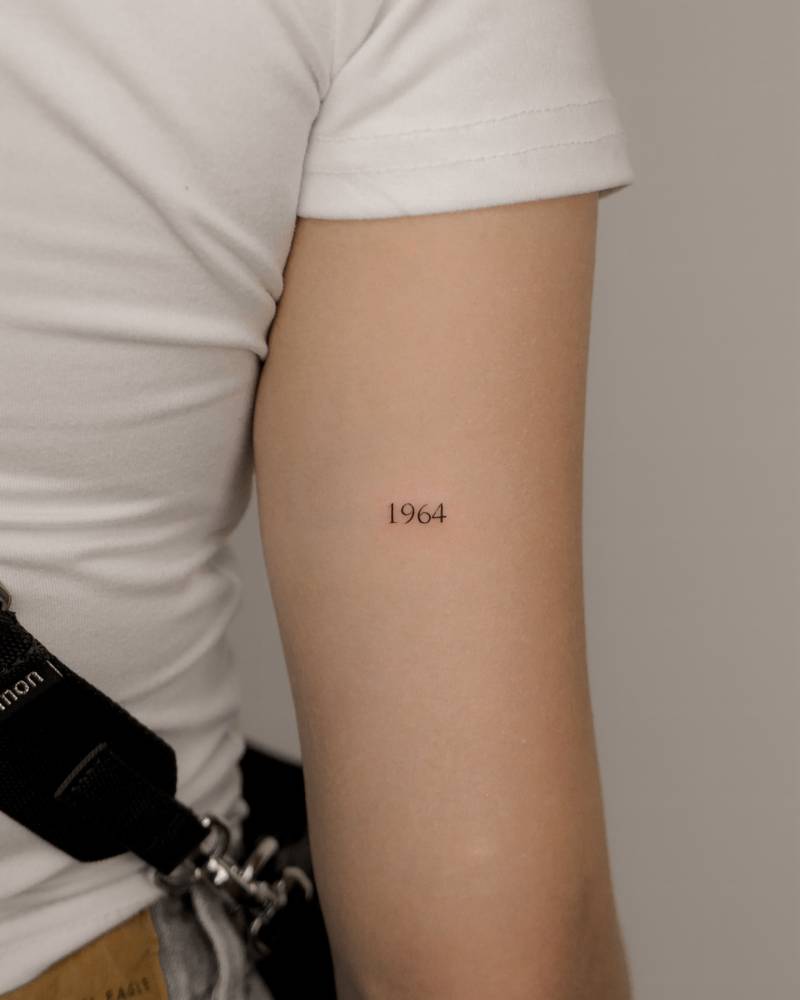 "1964" lettering tattoo in serif font located on the tricep.