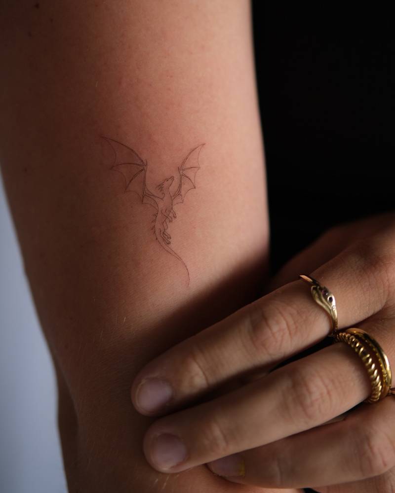 Tiny dragon tattoo in fine line style located on the upper arm.