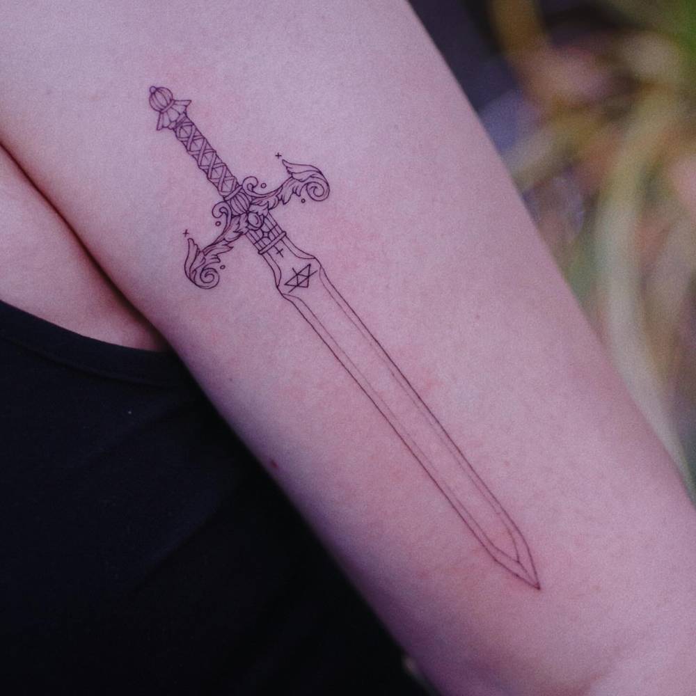 Fine line style sword tattoo located on the upper arm.