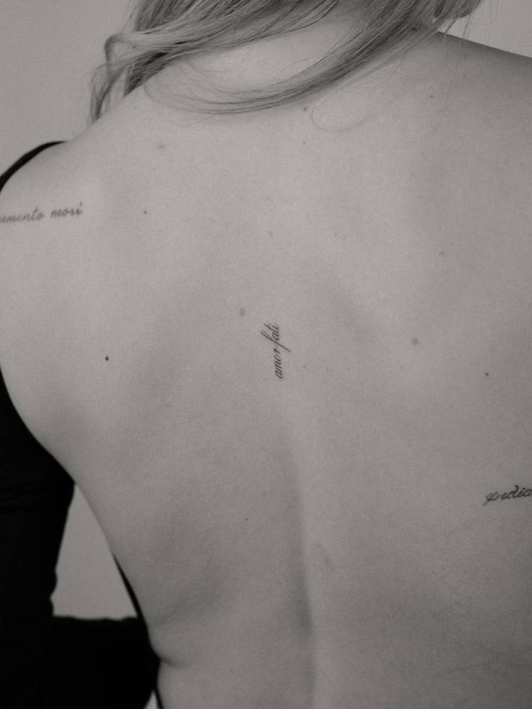 Script font "amor fati" lettering tattoo located on the upper back.