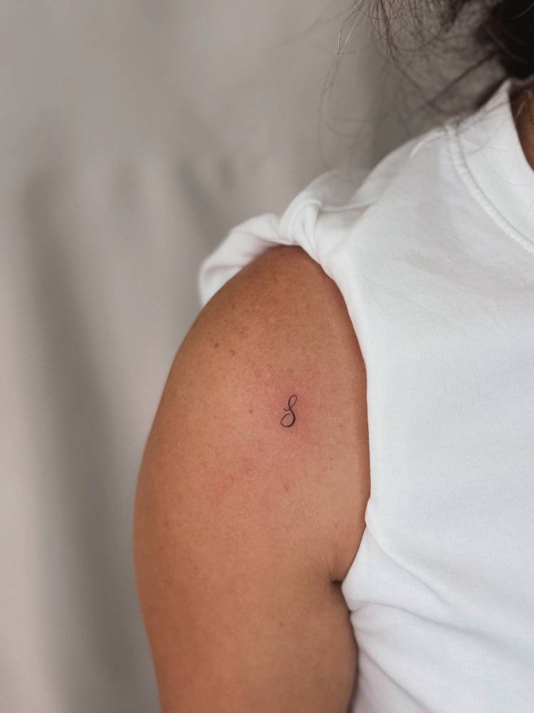 Script font letter "S" tattoo located on the shoulder.