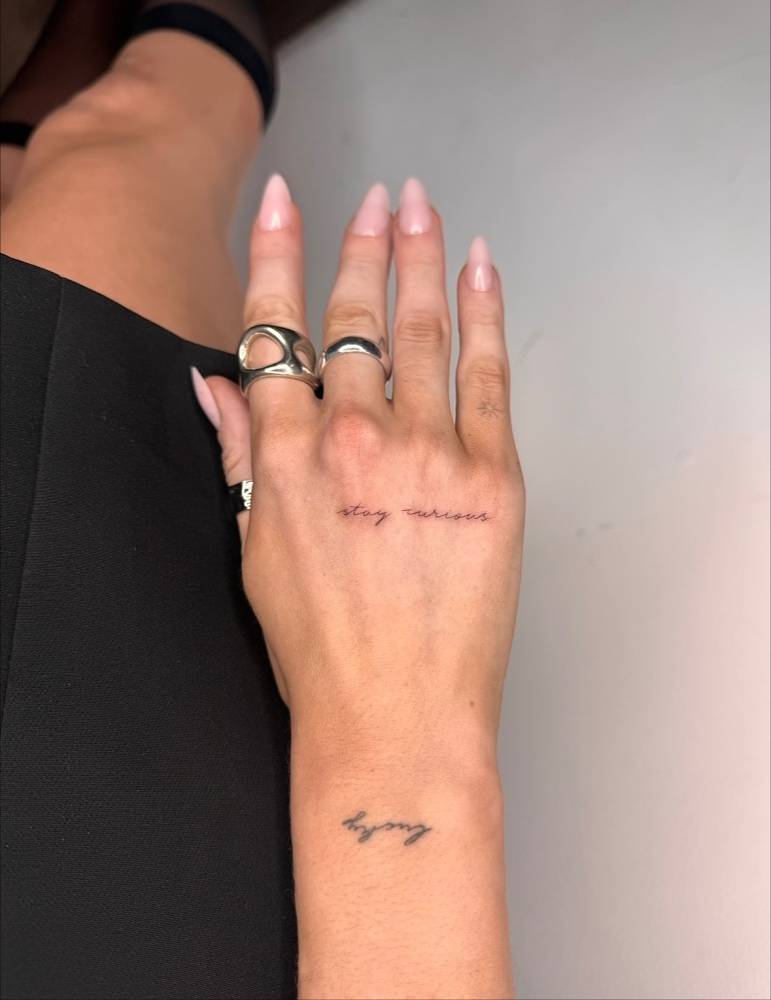 Handwritten font "stay curious" lettering tattoo located on the hand.
