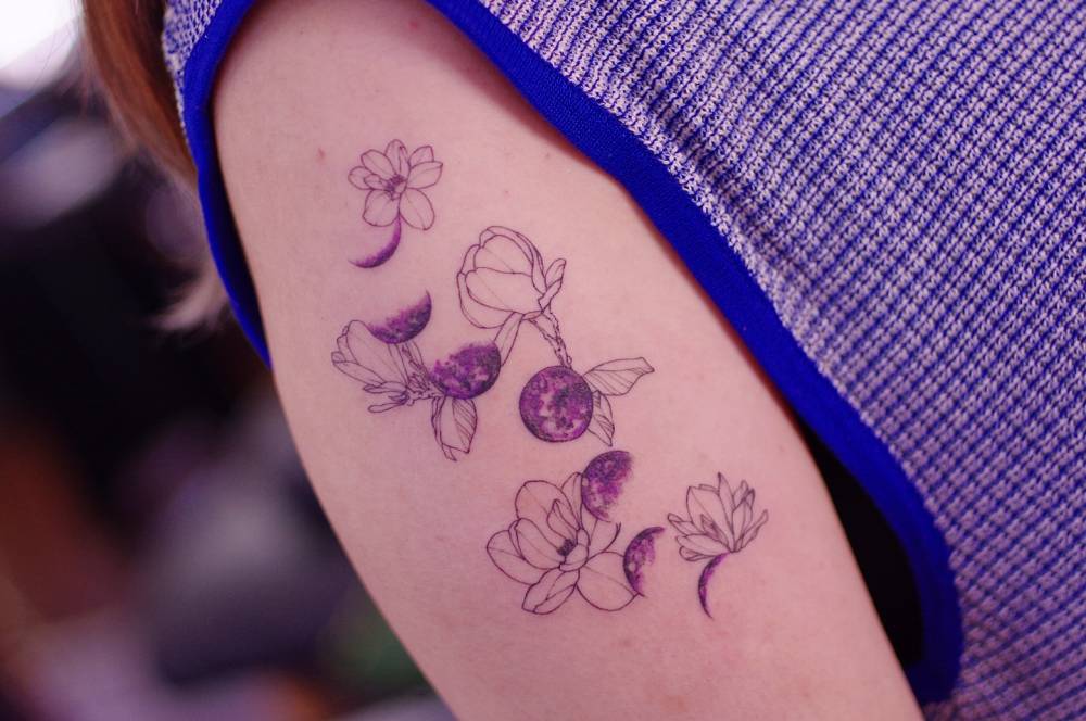 Moon phases and flowers tattooed on the upper arm, illustrative style.
