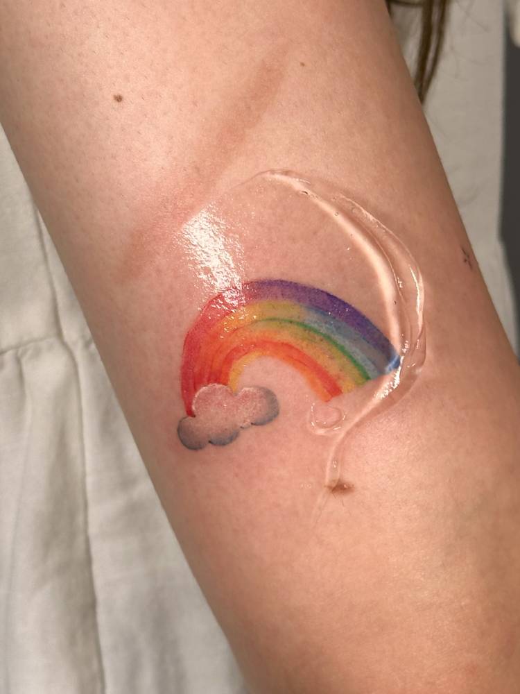 Watercolor style rainbow and cloud tattoo located on the upper arm.