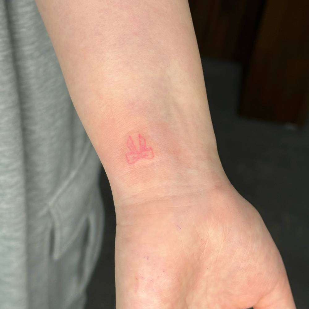 Minimalistic pink bow tattoo located on the wrist.