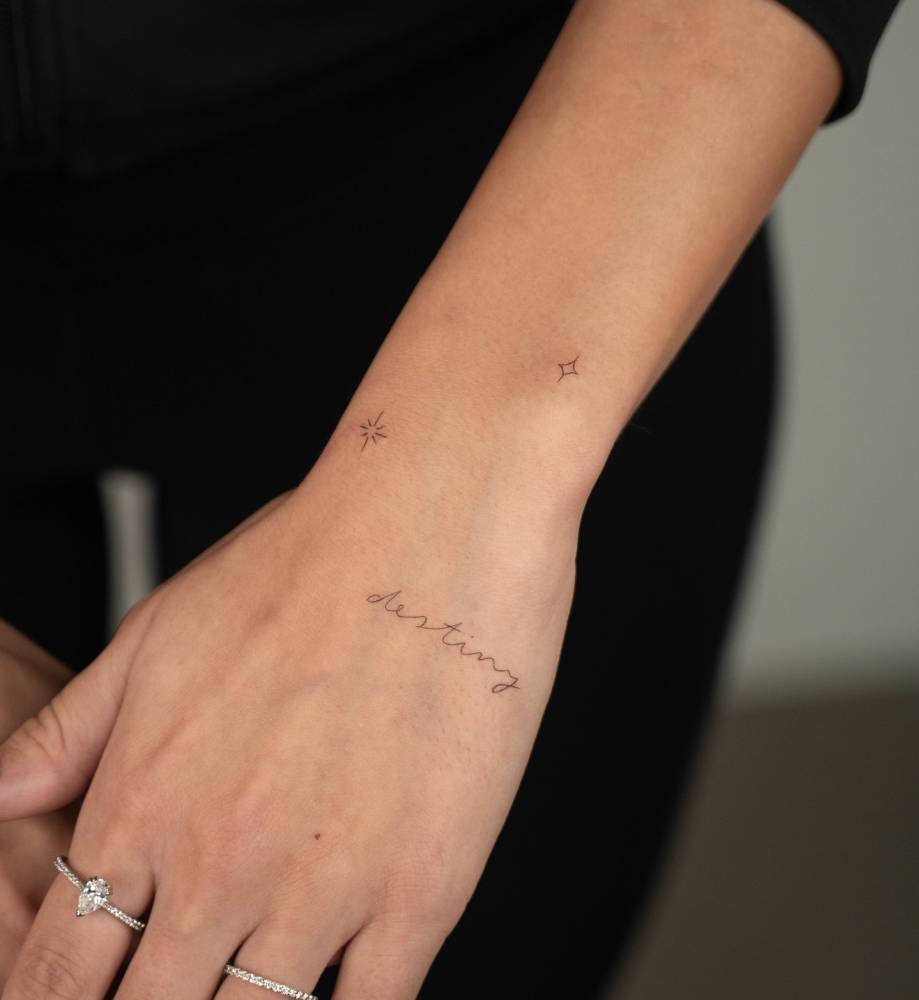 Word "destiny" and stars tattoo located on the hand and wrist.