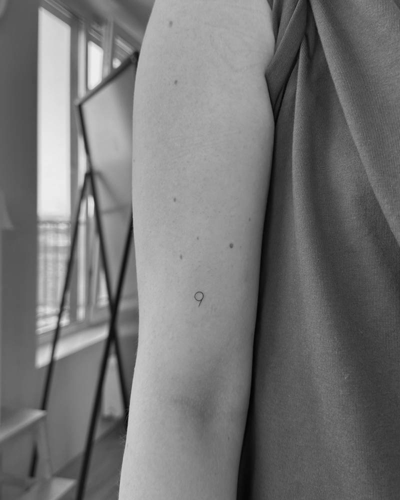 Number "9" tattooed on the bicep, done in minimalistic style.