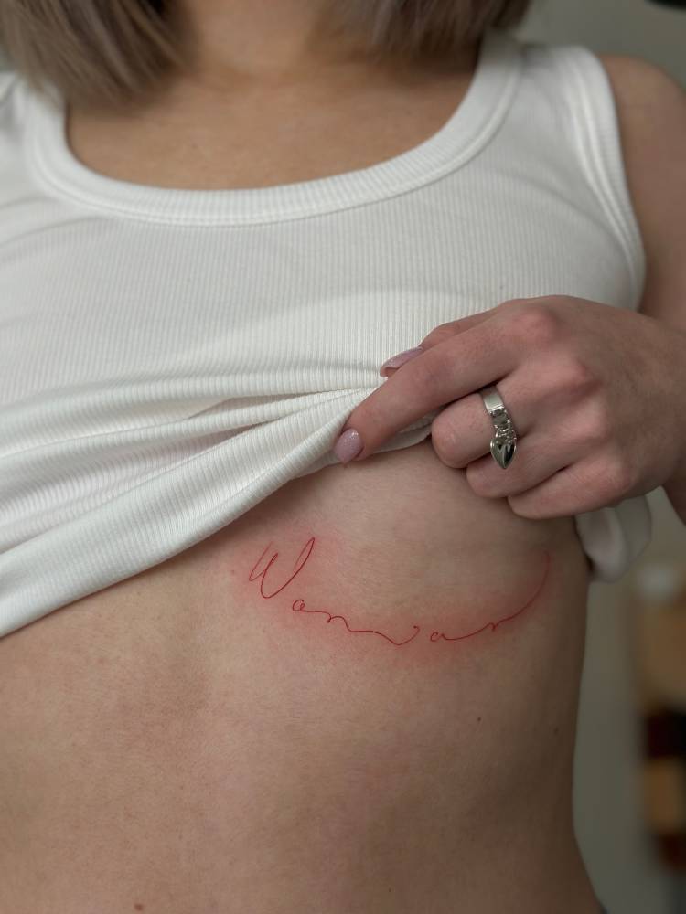 Handwritten font word "woman" in red ink tattooed on the under boob.