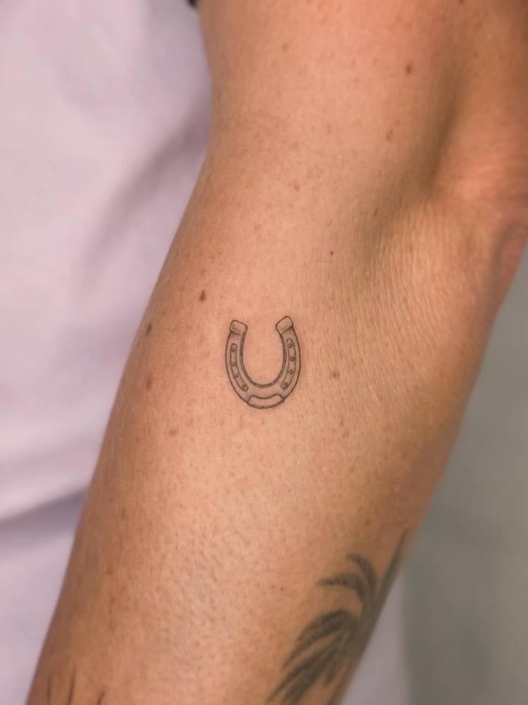 Tiny horseshoe tattoo in illustrative style located on the forearm.