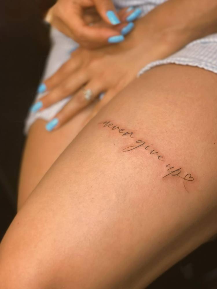 "Never give up" lettering tattoo located on the thigh.