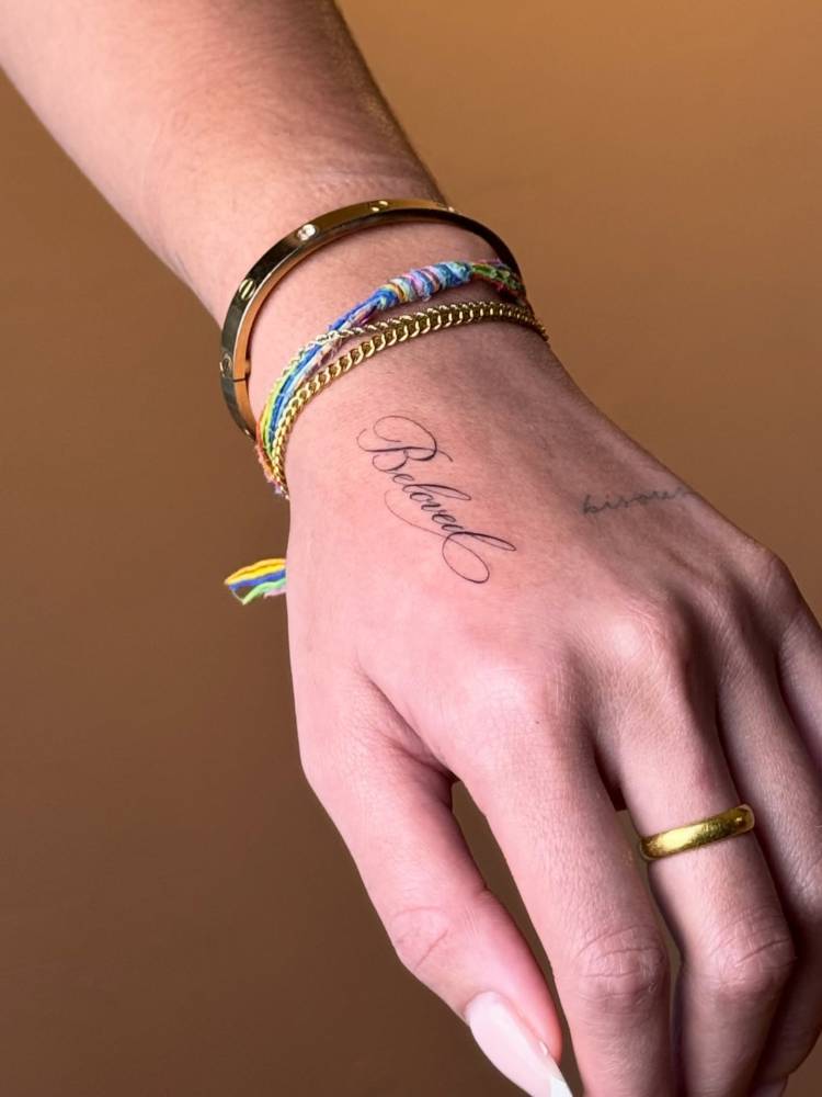 Script font word "beloved" tattooed on the hand.