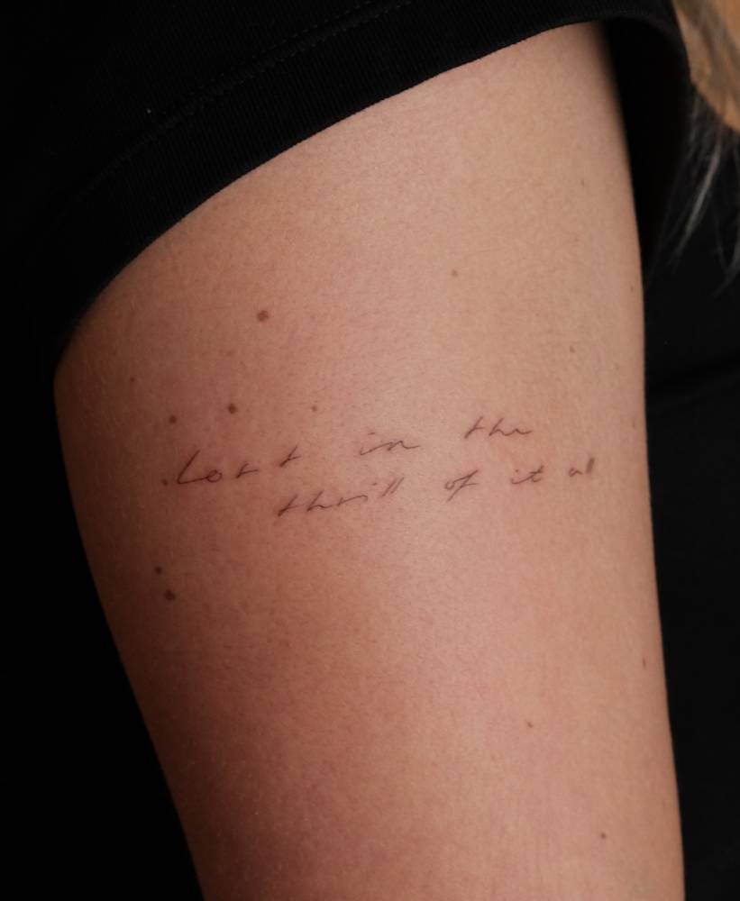 "Lost in the thrill of it all" lettering tattoo handwritten on the upper arm.