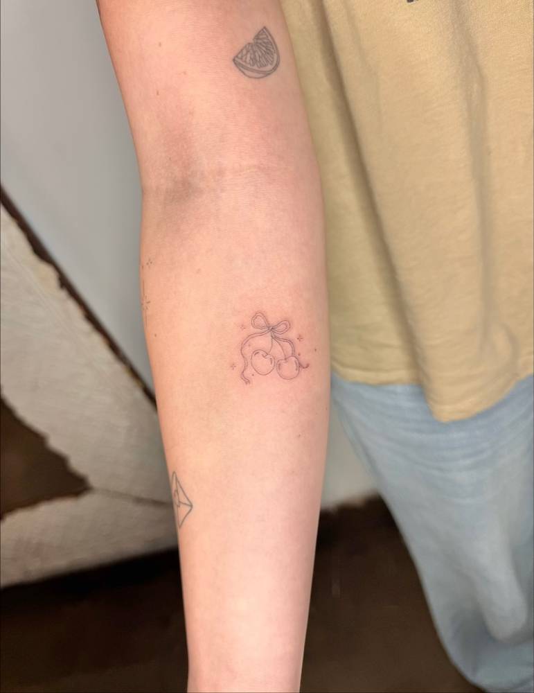 Cherries and bow tattoo in fine line style located on the inner forearm.