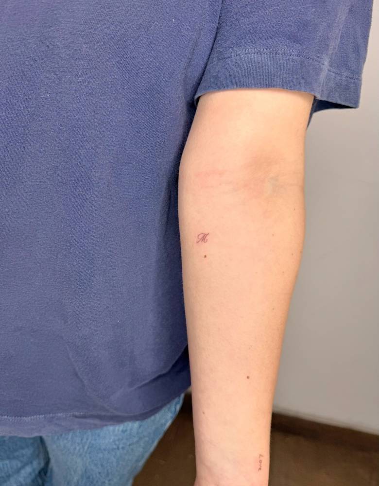 Script font tiny letter "M" tattoo located on the inner forearm.