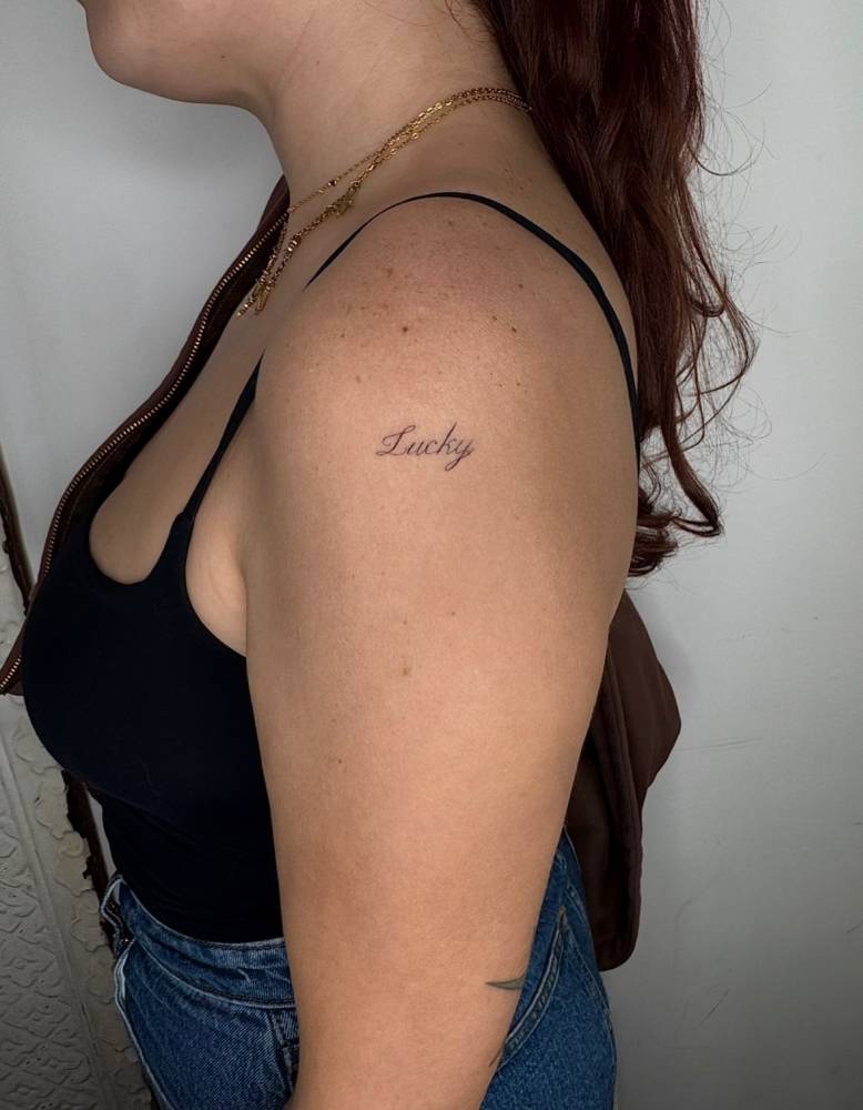Lettering tattoo of the word "lucky" located on the upper arm.