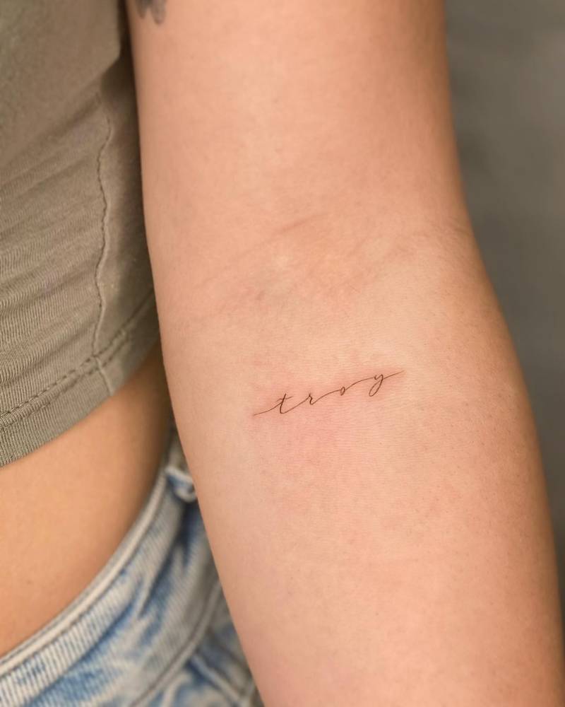 Name "Troy" tattoo in handwritten font located on the inner forearm.