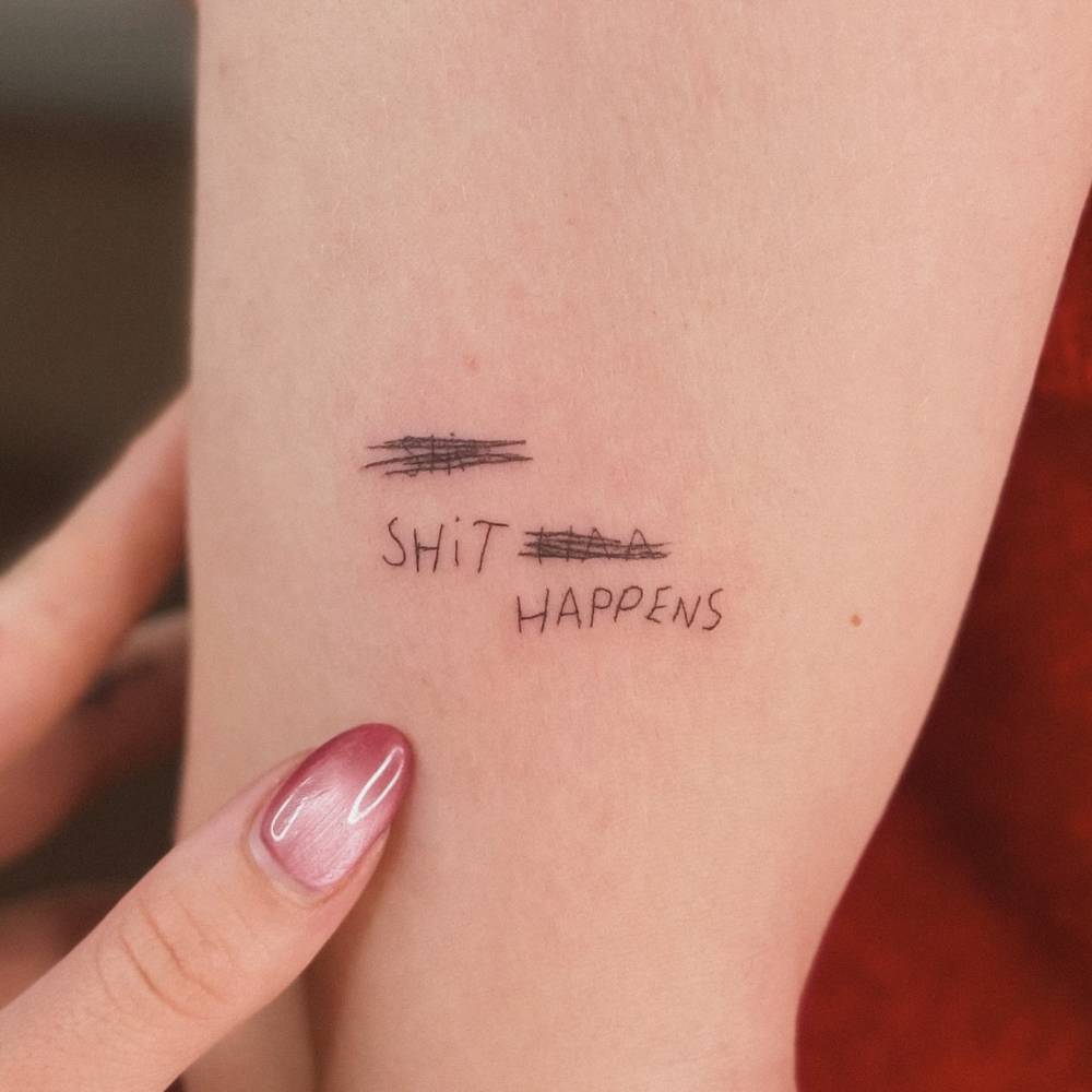 Handwritten font "shit happens" tattoo located on the upper arm.