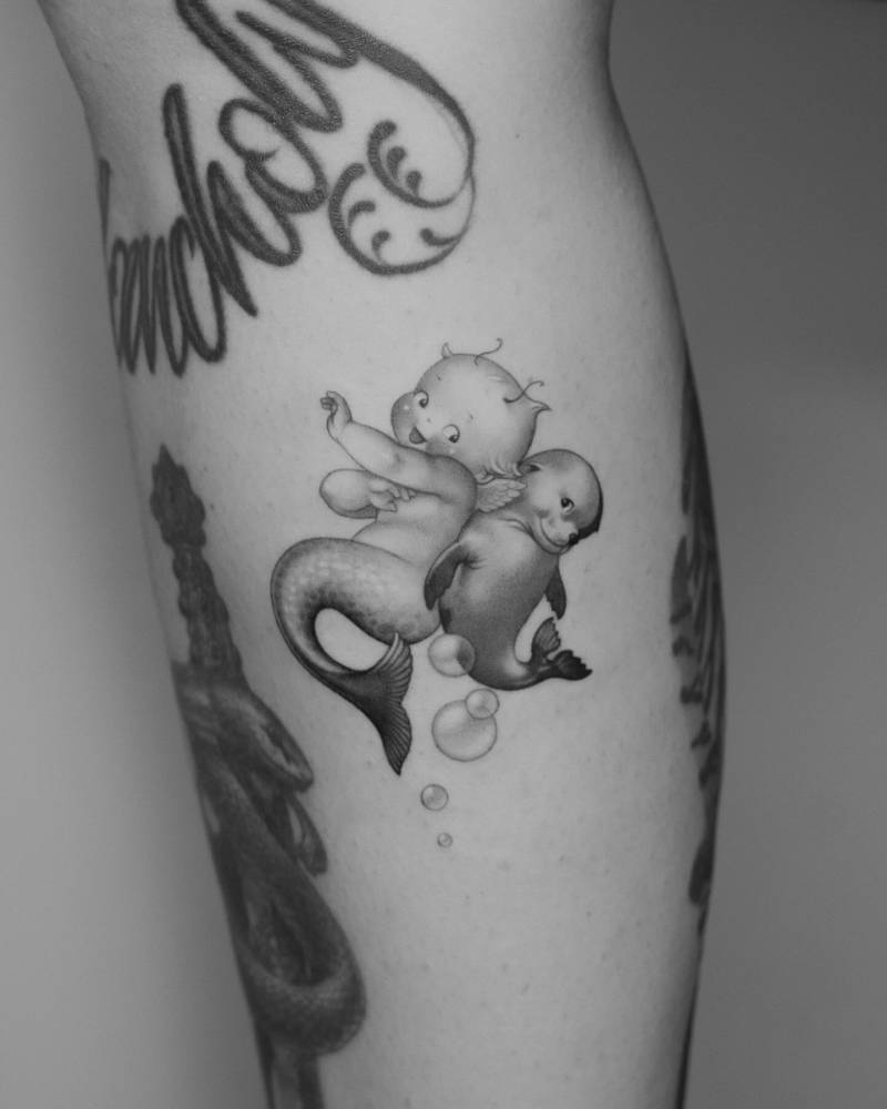 Kewpie mermaid and seal tattoo located on the calf, cartoon style.