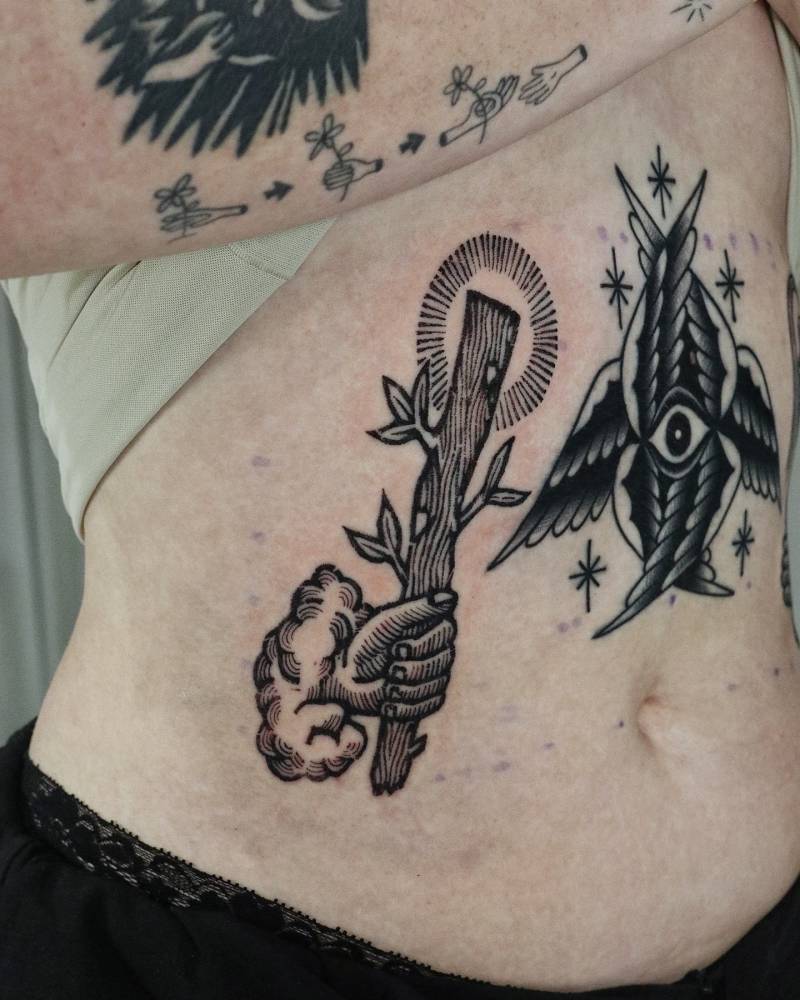 Hand holding a twig tattooed on the waist, engraving style.