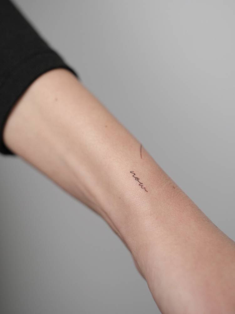 Handwritten font word "now" tattooed on the wrist.