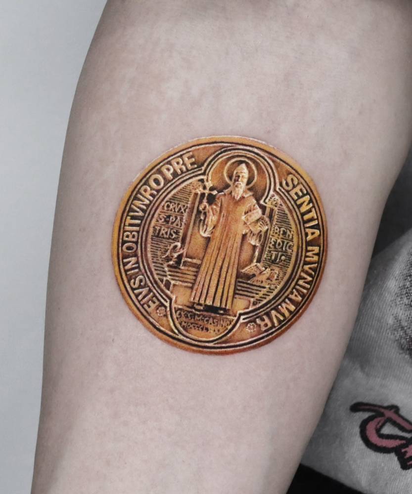 Micro-realistic style Saint Benedict medal tattooed on the inner forearm.