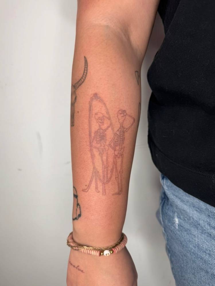 Skeleton looking in the mirror tattooed on the forearm, fine line style.