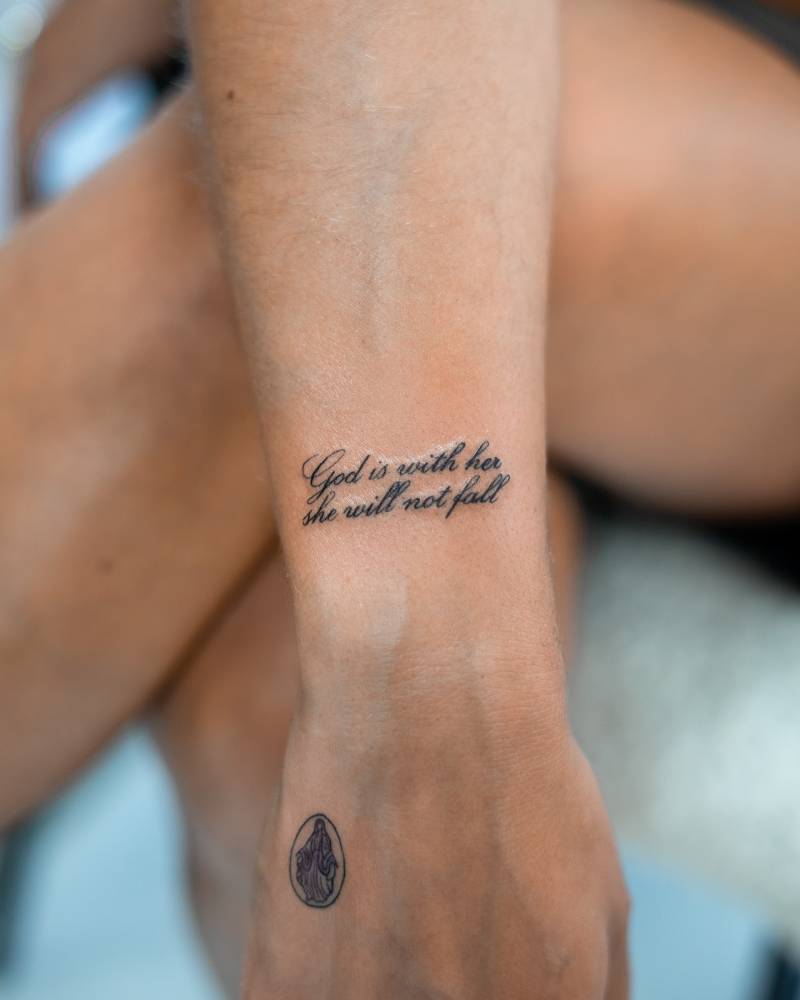 Script font "god is with her, she will not fall" tattoo located on the wrist.