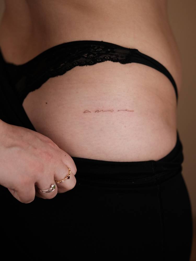 Handwritten font word "amore" tattooed on the hip.
