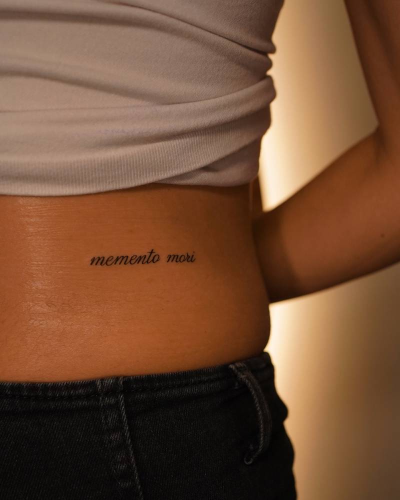 "Memento mori" lettering tattoo located on the lower back.