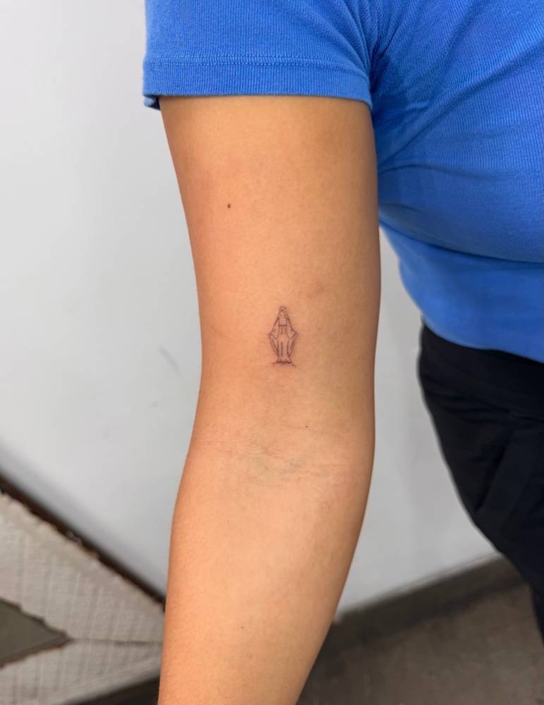 Tiny Virgin Mary tattoo located on the bicep, fine line style.