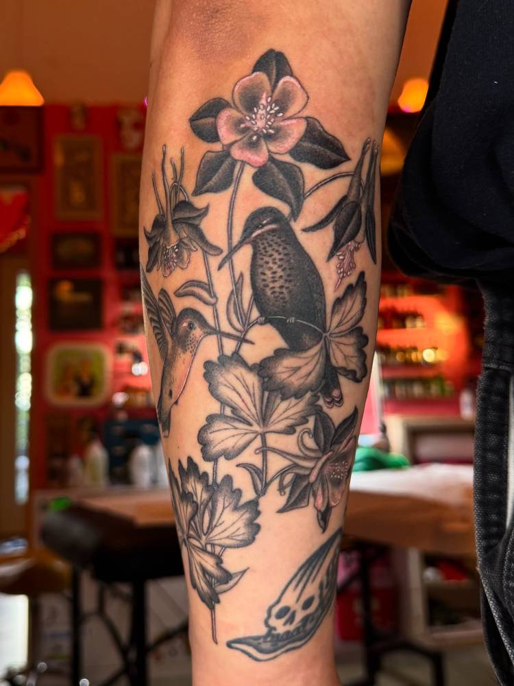 Illustrative style hummingbirds and flowers tattoo located on the forearm.
